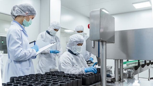 Clean room technicians monitor vial filling operation. Quality control inspector and factory workers operating automated packaging machinery in a sterile pharmaceutical manufacturing facility.