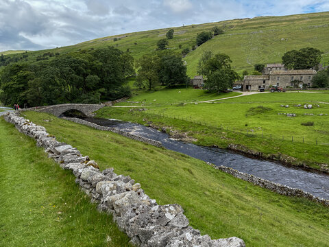 View of a historic stone bridge crossing a river in the Yorkshire Dales surrounded by lush green hills, stone walls, and traditional farm buildings Hawes, England, United Kingdom.