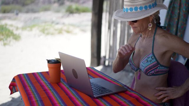 Mature woman shows emotion while using computer and tablet on a tropical beach while drinking coffee during vacation.