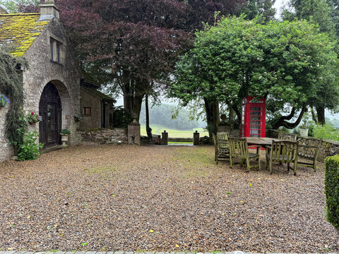 View of a traditional stone cottage with a gravel courtyard, wooden furniture, and a red telephone box overlooking the rolling hills of Yorkshire in Hawes, England, United Kingdom.