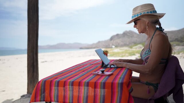 Mature woman shows concern while using computer and phone at beach. She enjoys her time working while on vacation.