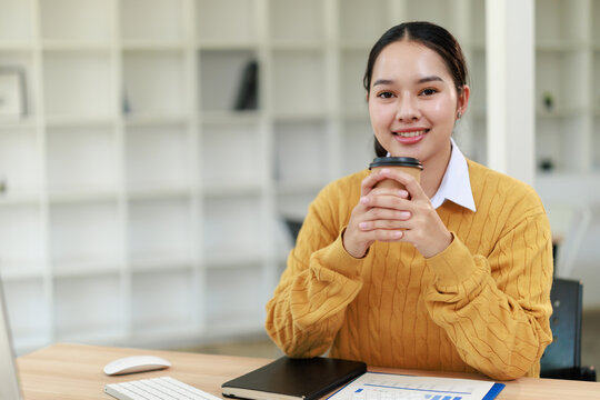 Asian woman smiling and holding a takeaway coffee cup, sitting at a desk with a notebook and documents, enjoying a relaxing break in a bright, modern office or educational environment