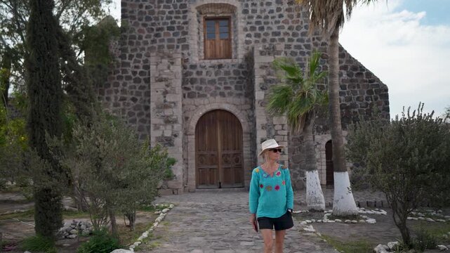 A mature woman strolls around Mission Church in Santa Rosalia, Mulege, with palm trees and mountains in Baja California Sur.