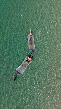 On a sunny day in Koh Kood Island, Thailand, a couple enjoys the tranquility of the turquoise sea while lounging on hammocks. Surrounded by lush greenery, this serene setting is perfect for unwinding.