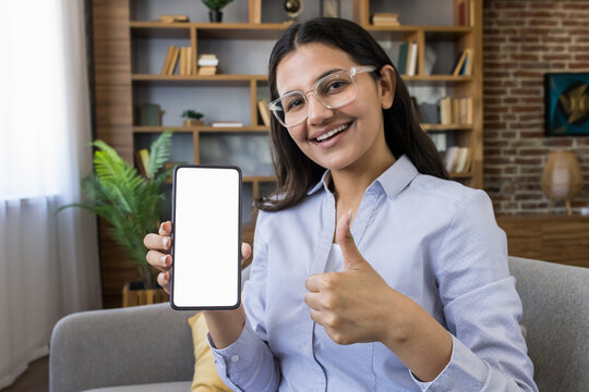 Young woman smiling and holding a smartphone with a blank white screen, giving a thumbs up gesture, recommending a new application or service from her home office setup