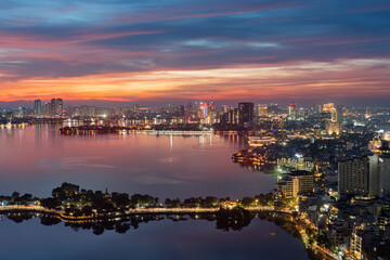 Fototapeta premium Evening aerial cityscape of West Lake and Thanh Nien Road featuring illuminated Hanoi skyline, reflective waters, and vibrant sunset colors.