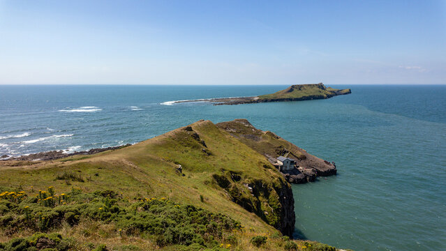 View of the grassy cliffs and the iconic Worm's Head headland stretching into the blue sea under a clear sky in Swansea, Wales, United Kingdom.
