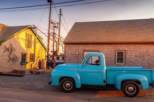 View of a vintage blue pickup truck with a dog in the driver's seat parked by shingled coastal buildings and fishing boat masts in Edgartown, Massachusetts, United States.