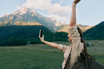 Fototapeta premium A woman outdoors takes a selfie with one arm raised, backpack on, against a mountain landscape. A lifestyle moment of exploration, freedom, and adventure in natural light.