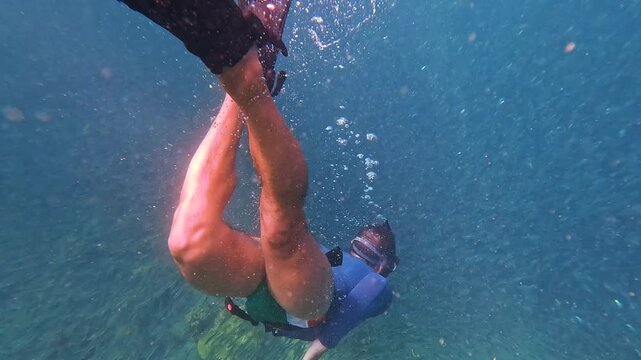 A person free dives deep into the ocean among an enormous school of fish. Bubbles rise as the diver swims close to the vast marine life