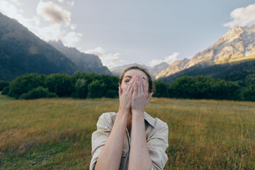 Fototapeta premium A young woman with brunette hair covers her face with her hands in a meadow as distant mountains rise behind her, expressing surprise and wonder.