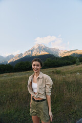 Young woman stands in an open meadow with a dramatic mountain backdrop, smiling softly under warm natural light, her relaxed stance suggesting carefree outdoor living