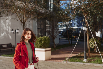 Fototapeta premium Young woman in a red coat walks thoughtfully along a city sidewalk on an autumn day with trees and street lamps in the background.