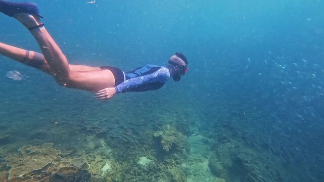 A person swims with snorkel gear and flippers above a beautiful coral reef in clear blue ocean water, passing a school of small fish during daylight