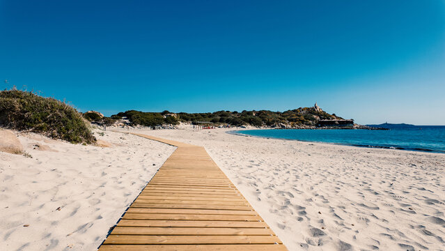 View of a wooden boardwalk leading across the white sand of Punta Molentis Beach towards the turquoise sea under a clear blue sky Villasimius, Sardinia, Italy.