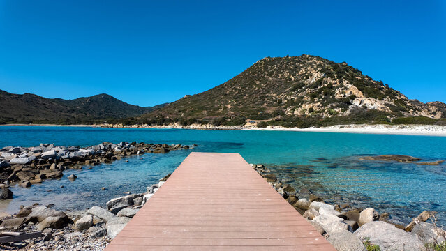 View of Punta Molentis Beach with a wooden pier leading into turquoise water, white sand, and rugged hills under a clear blue sky Villasimius, Sardinia, Italy.