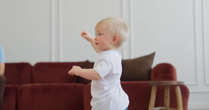 Blond toddler in a white shirt taking first steps across a bright living room with a red sofa and wooden stool in soft natural light