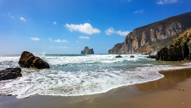 View of Scoglio Pan di zucchero sea stack from the sandy shore of Masua beach with crashing waves and rugged cliffs under a blue sky Masua, Sardegna, Italy.