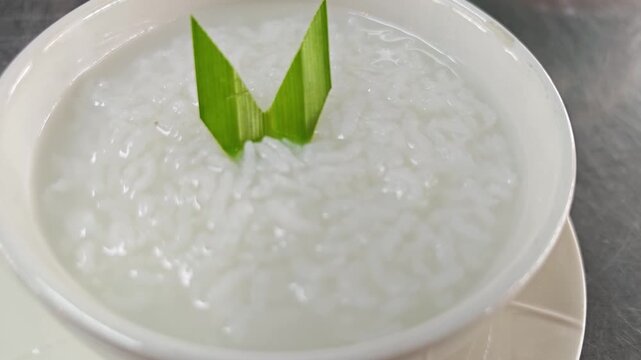 Close-up of hot white rice porridge decorated with pandan leaf in a ceramic bowl.