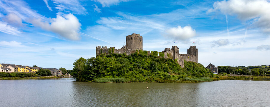 View of Pembroke Castle standing on a lush green hill overlooking the calm water under a blue sky with wispy clouds in Pembroke, Wales, United Kingdom.