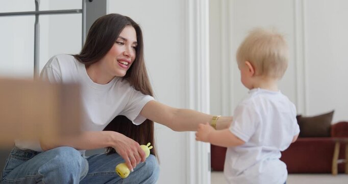 Young mother sitting on floor and encouraging blond toddler playing with a yellow toy microphone in bright living room during family bonding time