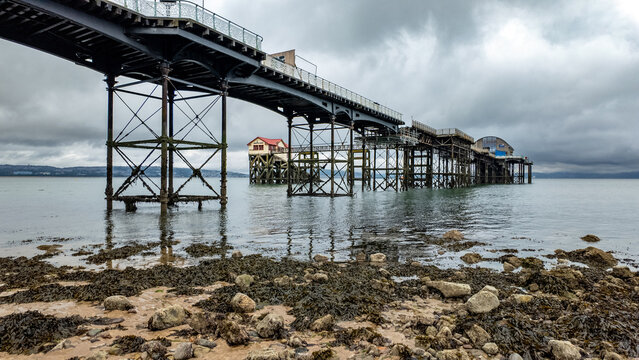 View of Mumbles Pier extending over the sea from a rocky shoreline under a dramatic overcast sky Mumbles, Wales, United Kingdom.