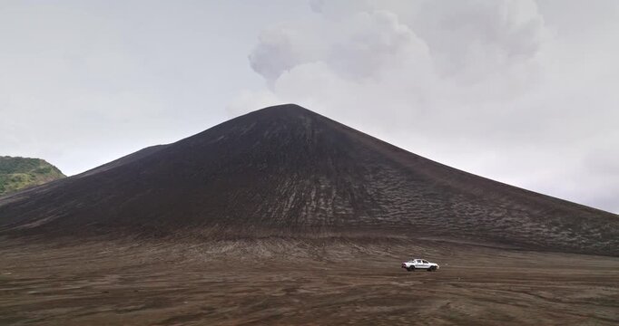 White pickup truck traversing the desolate volcanic ash plain, driving past the impressive Mount Yasur volcano under a grey sky, highlighting adventure travel and rugged exploration in Vanuatu