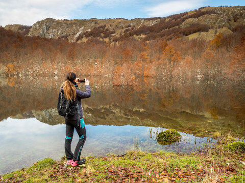 A female hiker photographs the scenery with a smartphone, capturing a travel adventure by an ephemeral lake, Resia Valley, Julian Alps, Friuli Venezia Giulia, Italy.