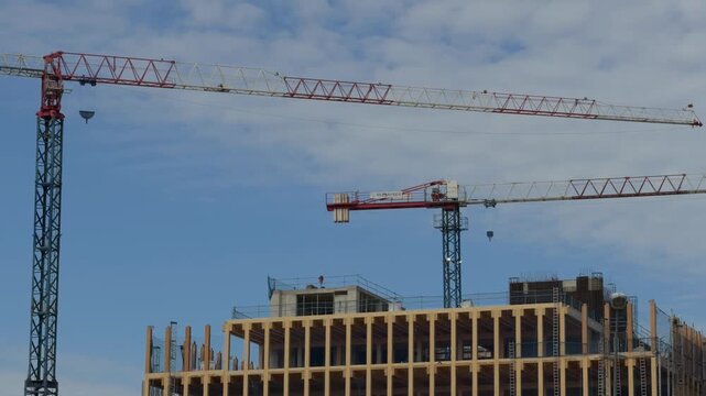 Tower cranes moving on a construction site while erecting a modern sustainable wooden building against a partly cloudy blue sky, showcasing real estate development and urban progress