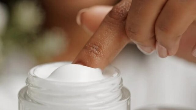 Close up motion shot of finger dipping into luxurious white cream in clear glass jar, skincare product for beauty and self care routine.