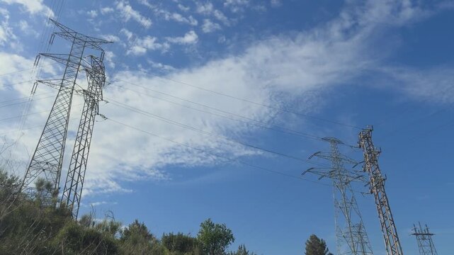 High voltage power lines and transmission towers under a blue sky with moving white clouds representing energy grid infrastructure and electricity distribution