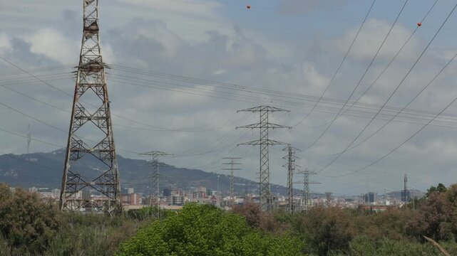 High voltage power lines and transmission towers carrying electricity over a natural landscape with a modern city skyline against a cloudy sky on a dull day