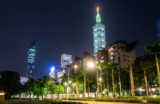 Taipei, Taiwan - April 16, 2025: Glowing bamboo-inspired Taipei 101 tower rises with nearby high rises above a palm lined park and boulevard on a clear night.