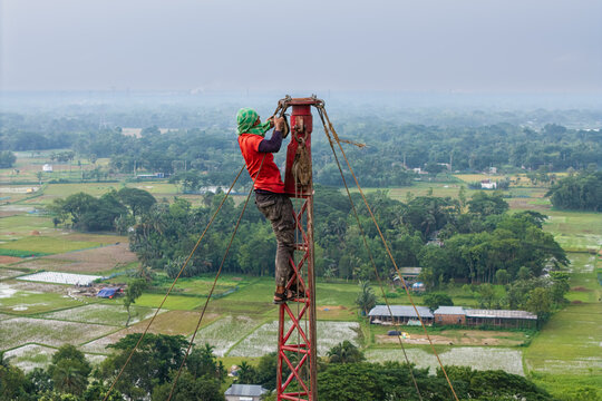 Trishal Upazila, Bangladesh - 13 May 2026: Aerial view of a worker climbing a red metal tower using ropes and pulleys over a lush green rural landscape with agricultural fields.