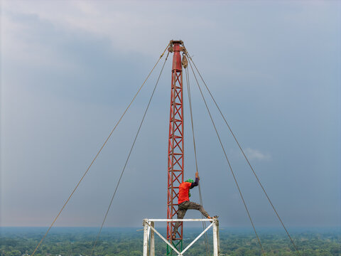 Trishal Upazila, Bangladesh - 13 May 2026: Aerial view of a worker climbing a tall red lattice mast with guy wires against a vast green landscape and cloudy sky.