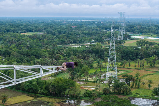 Trishal Upazila, Bangladesh - 13 May 2026: Aerial view of a man lying on the arm of a high-voltage electricity pylon overlooking a lush green landscape with fields and trees.
