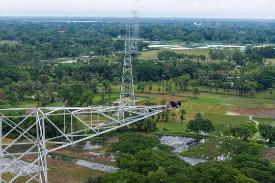 Trishal Upazila, Bangladesh - 13 May 2026: Aerial view of a worker sitting on a high-voltage electricity pylon overlooking a lush green rural landscape with fields and trees.