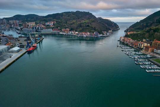 Pasai Donibane, Spain - 10 March 2026: Aerial view of the Harbour mouth of Pasaia with its narrow channel, coastal buildings, and lush green hills under a cloudy sky.