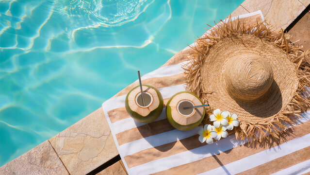 Two coconut drinks with straws beside straw hat on striped pool towel. Tropical resort refreshment. Luxury summer vacation flat lay. Poolside travel promo.