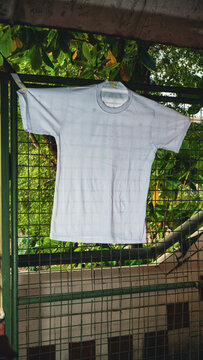 White t-shirt hangs on a metal fence. Green leaves frame the top of the shot. Background shows distant buildings and trees. Soft daylight casts gentle shadows. Image suggests a quiet, everyday moment
