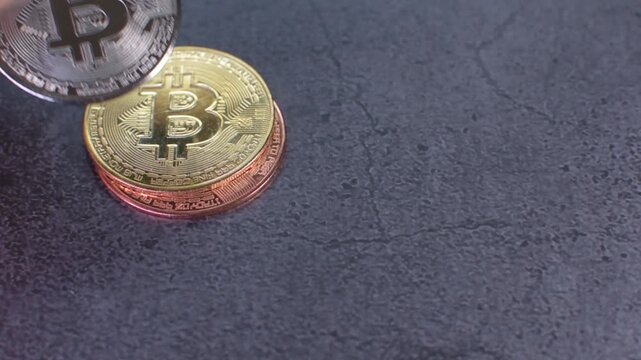 Video of a hand entering the frame to pick up a silver bitcoin cryptocurrency coin from stacked gold and copper coins placed on a dark gray granite surface, representing blockchain technology, digital