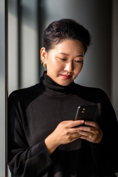 Middle-aged Asian female architect with fair skin and a plus size body checks her smartphone while wearing a black sweater.