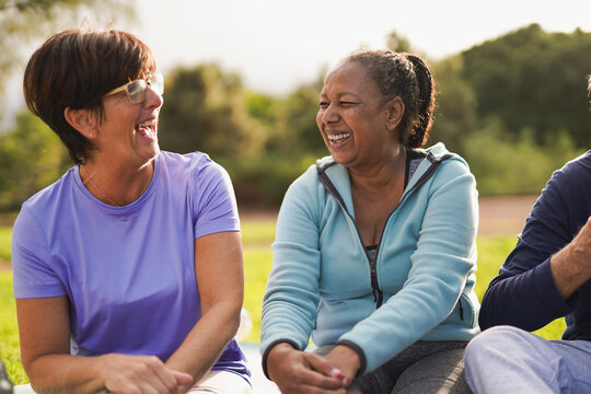 Happy multiracial senior people having fun together sitting at city park after yoga class during sunset time - Healthy elderly friends, diversity and fit lifestyle concept