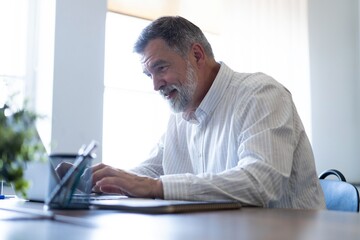 Obraz premium Smiling mature business man executive wearing shirt sitting at desk using laptop. Happy busy professional middle aged businessman investor working on computer looking away in office