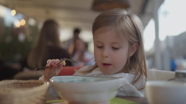 A happy, funny, smiling little girl cheerfully eats food in a restaurant. A child eats food with gusto on a street cafe terrace. Kid child holding fork in hand.