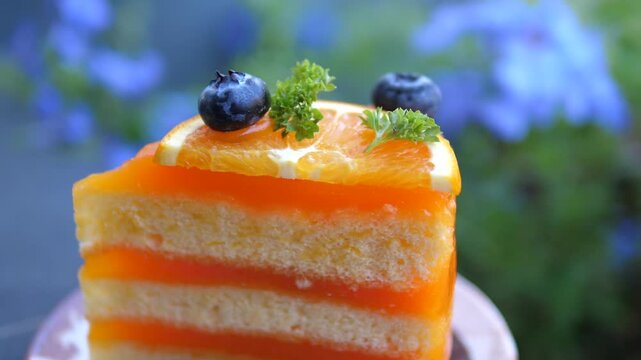 Cinematic close-up of a delicious slice of layered orange sponge cake topped with fresh orange slice and blueberries, blurred blue flowers background