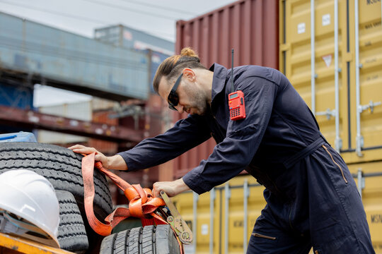 Worker in coveralls securing cargo straps around stacked tires beside shipping containers at a port freight yard.
