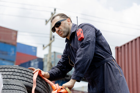 Worker in coveralls and sunglasses securing tires with straps at a shipping yard with stacked containers.