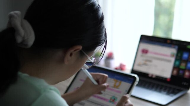Cinematic over the shoulder shot of a young female professional with glasses holding a stylus pen, looking at blurred multi-screen laptop and tablet software layout on desk