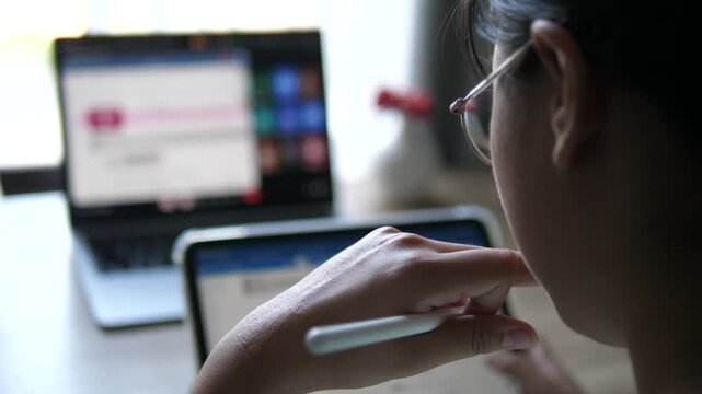Cinematic over the shoulder shot of a young female professional with glasses holding a stylus pen, looking at blurred multi-screen laptop and tablet software layout on desk